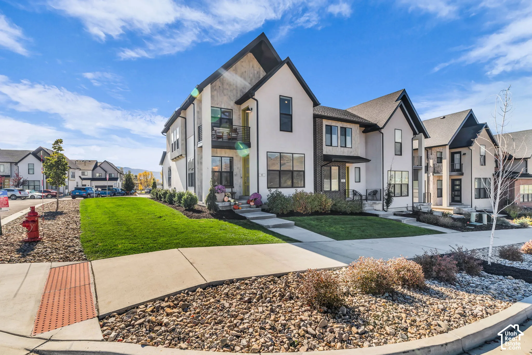 Multi-story exterior featuring light-toned stucco and dark brick accents, high-pitched rooflines, integrated private balcony with metal railing, and concrete walkways