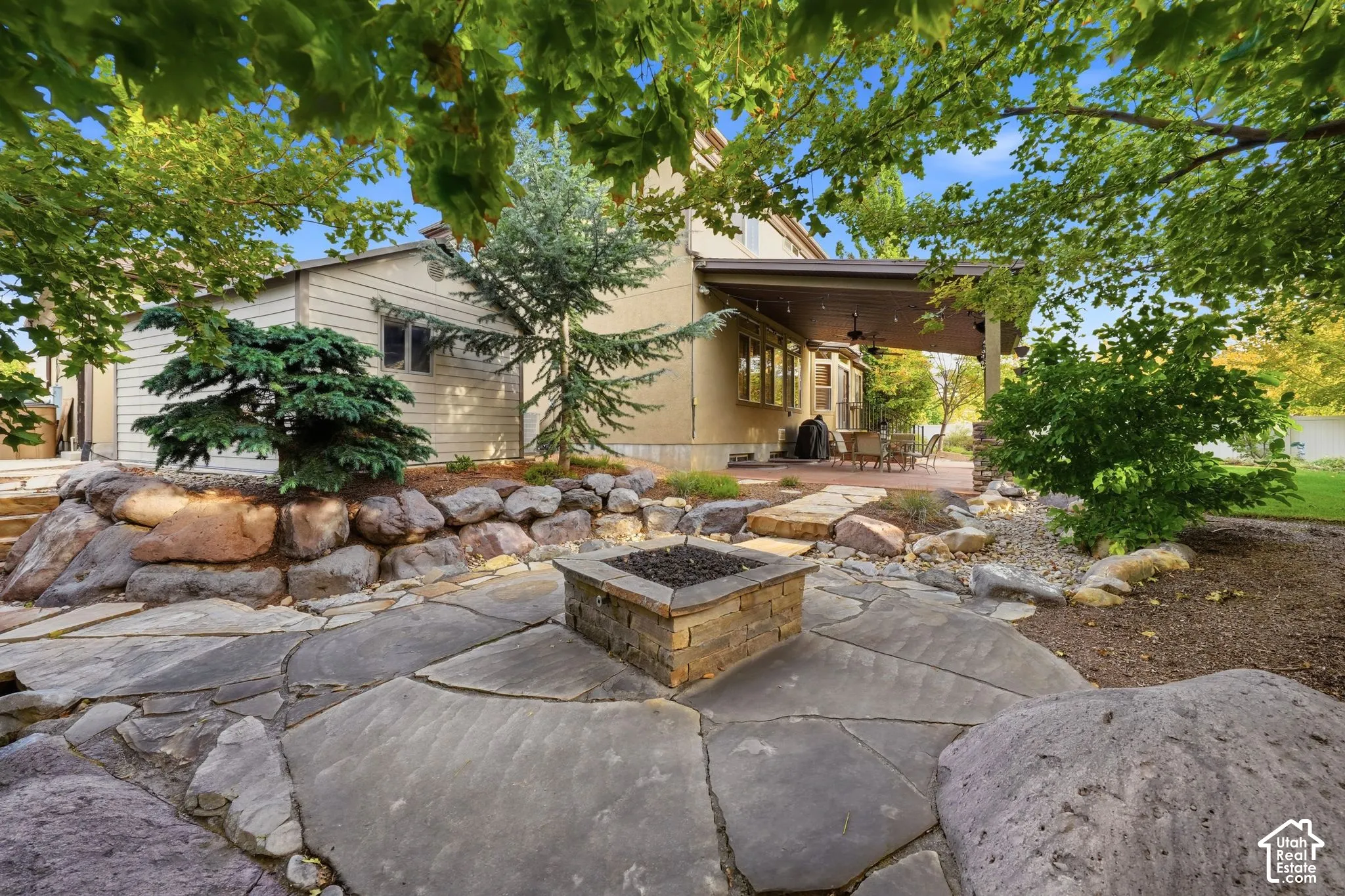 Backyard area featuring a stone patio, built-in stone fire pit, tiered boulder retaining wall, and a covered patio with recessed lighting