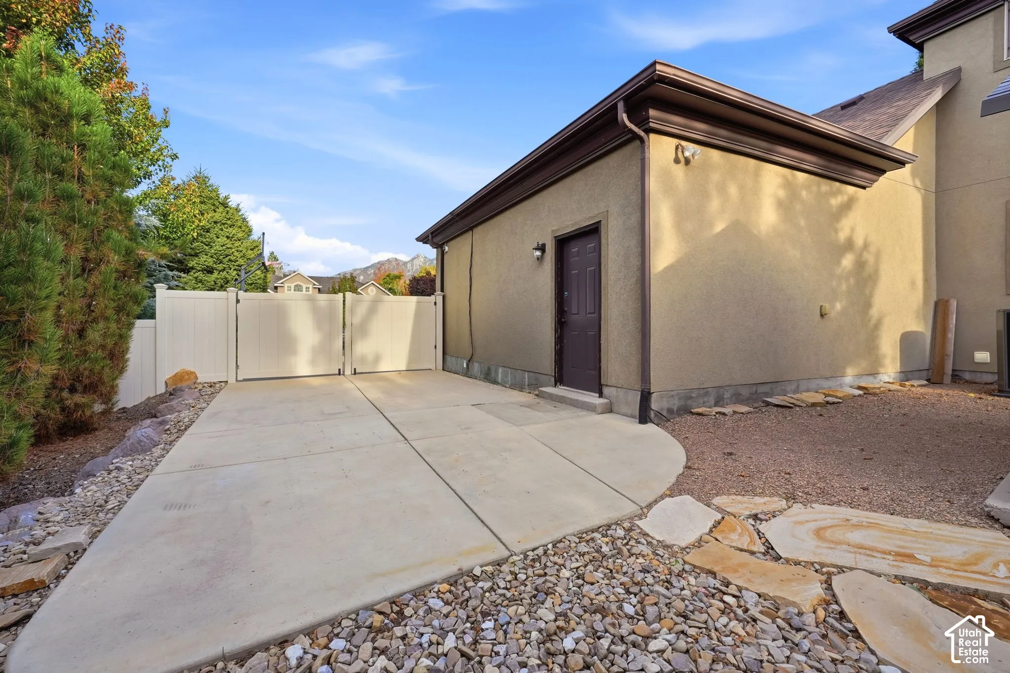 Stucco exterior featuring a concrete parking pad, gated side yard access, stone-paved landscaping, and exterior wall-mounted lighting