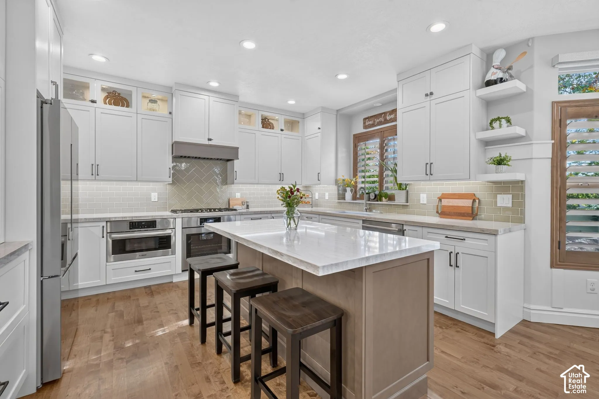 Kitchen featuring white shaker-style cabinetry, a central island with a stone countertop, herringbone-patterned backsplash, stainless steel appliances, and wood-finish flooring