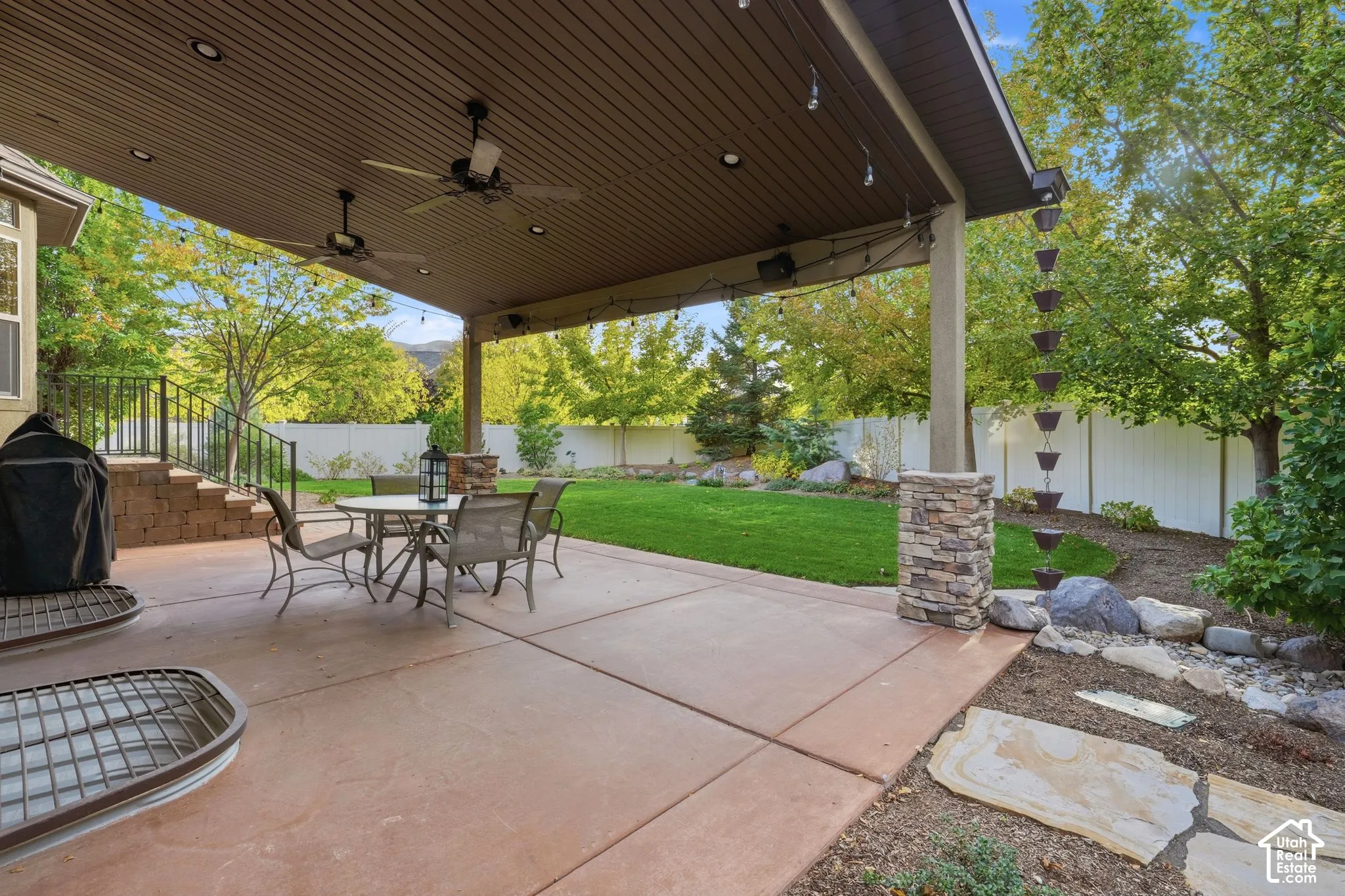 Covered concrete patio featuring recessed lighting, ceiling fans, stone-base columns, and a vertical metal rain chain