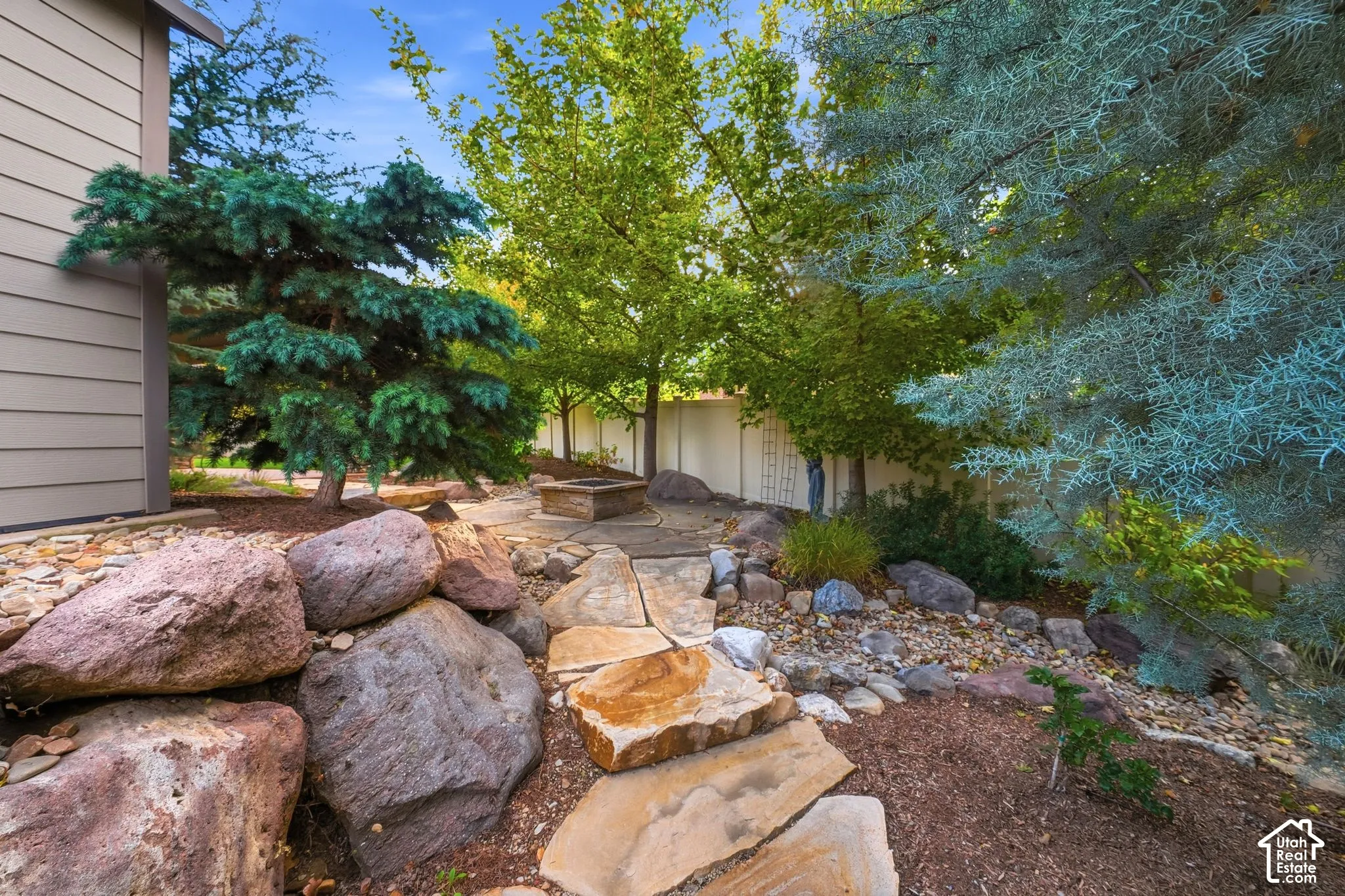 Backyard area featuring a stone flagstone path, built-in stone fire pit, rock landscaping, and mature trees