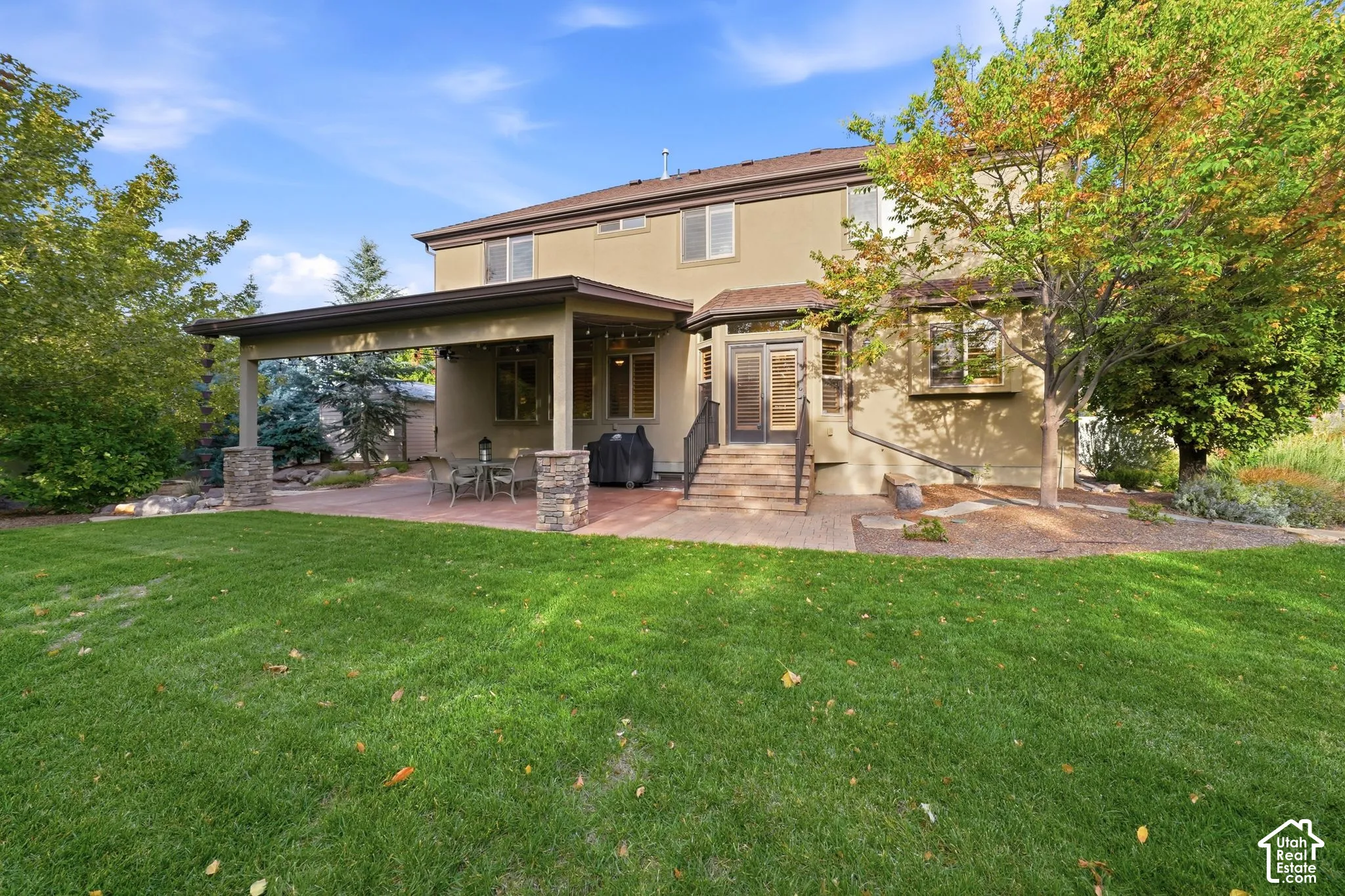 Stucco exterior featuring a covered patio, stone-base columns, paver-style hardscaping, and an expansive lawn with mature landscaping
