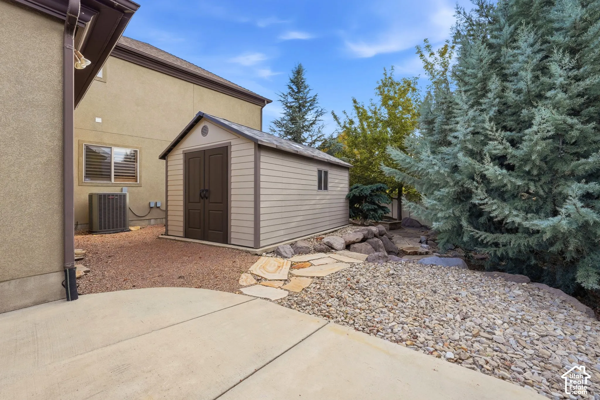 Exterior storage shed featuring a neutral siding finish, concrete patio, stone walkway, and rock landscaping
