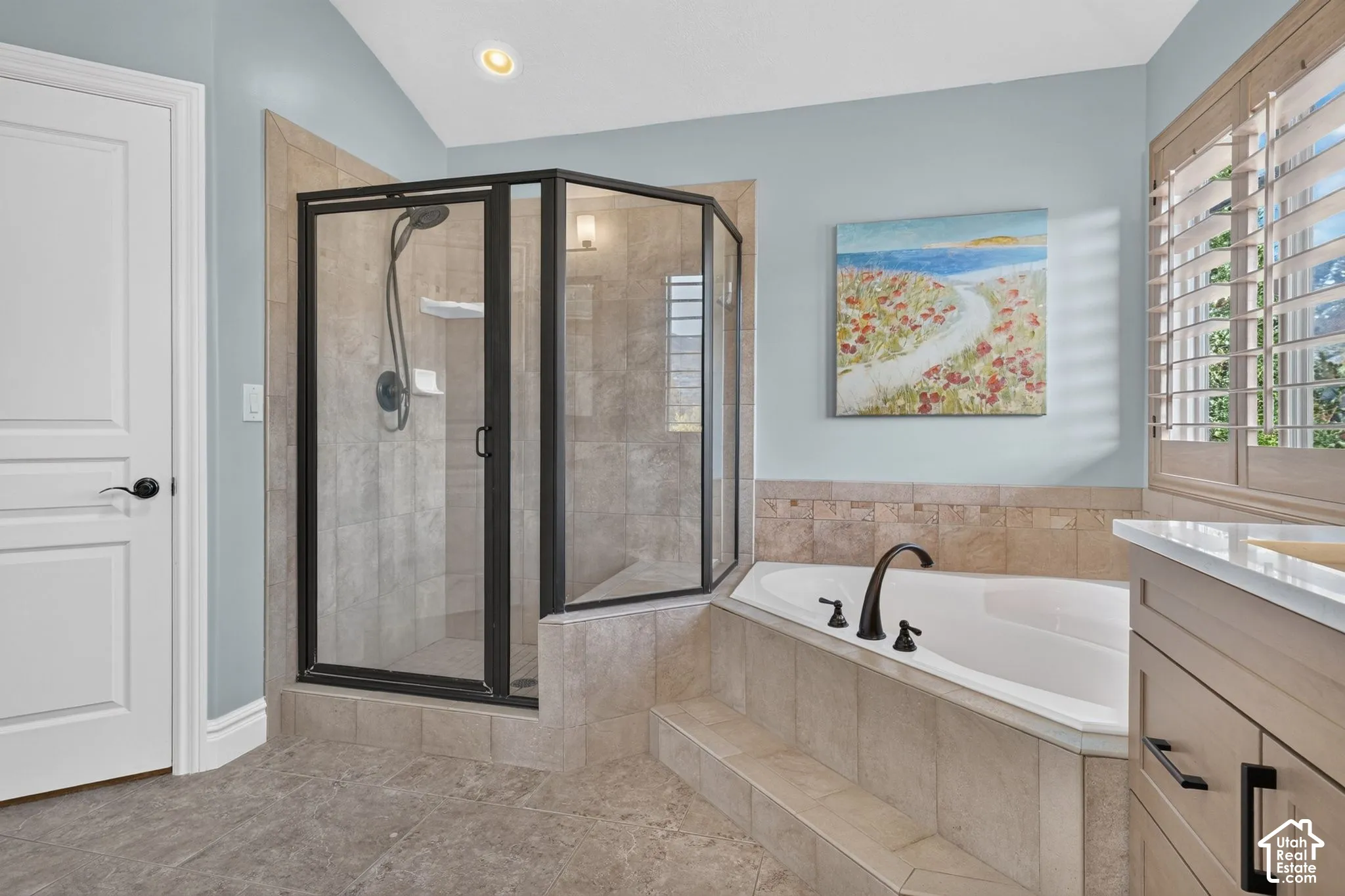 Corner soaking tub with tile surround, glass-enclosed shower with matte black fixtures, and light-toned wood cabinetry