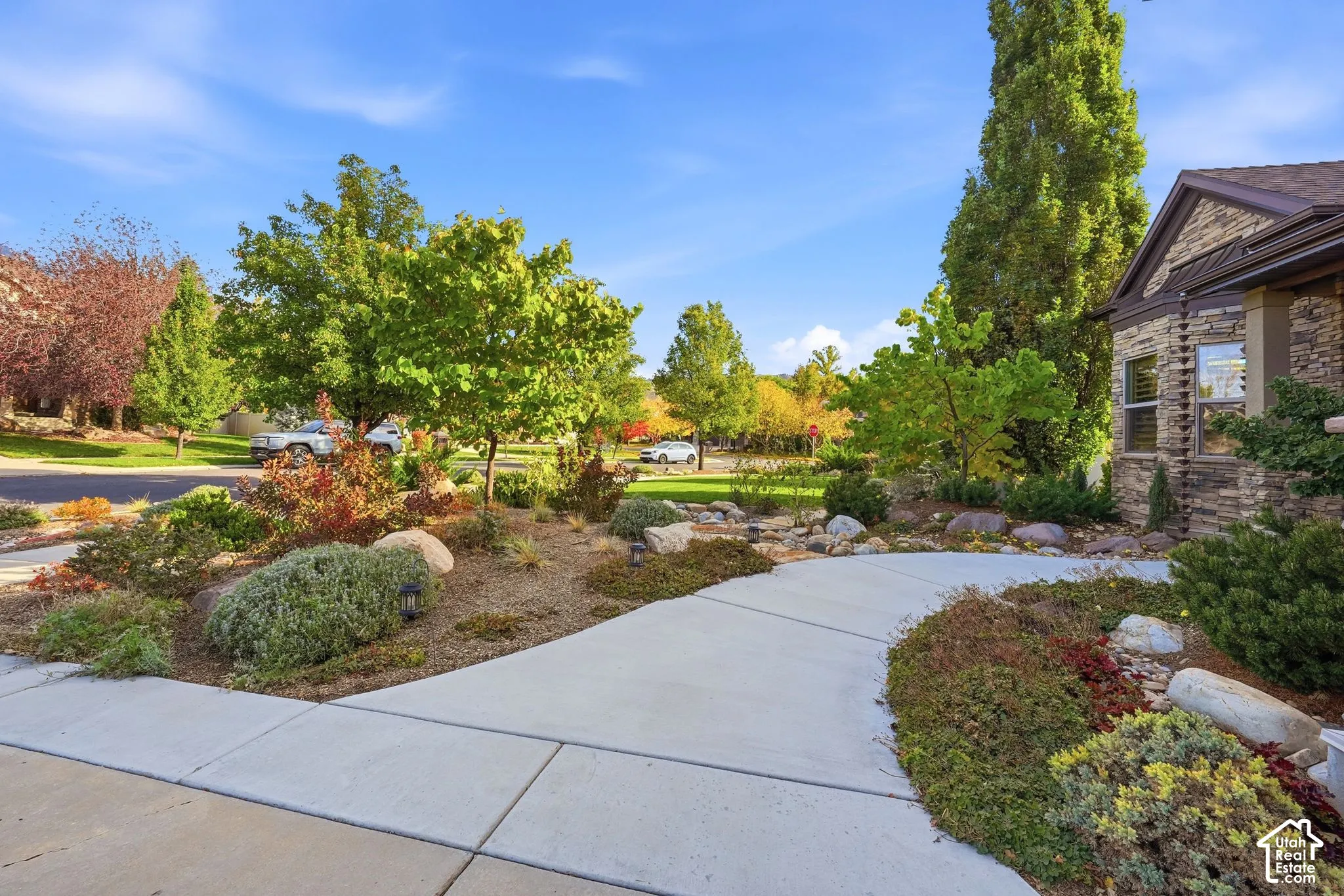 Front exterior featuring stone masonry cladding, a concrete walkway, and landscaped garden beds with mature trees and decorative rock elements