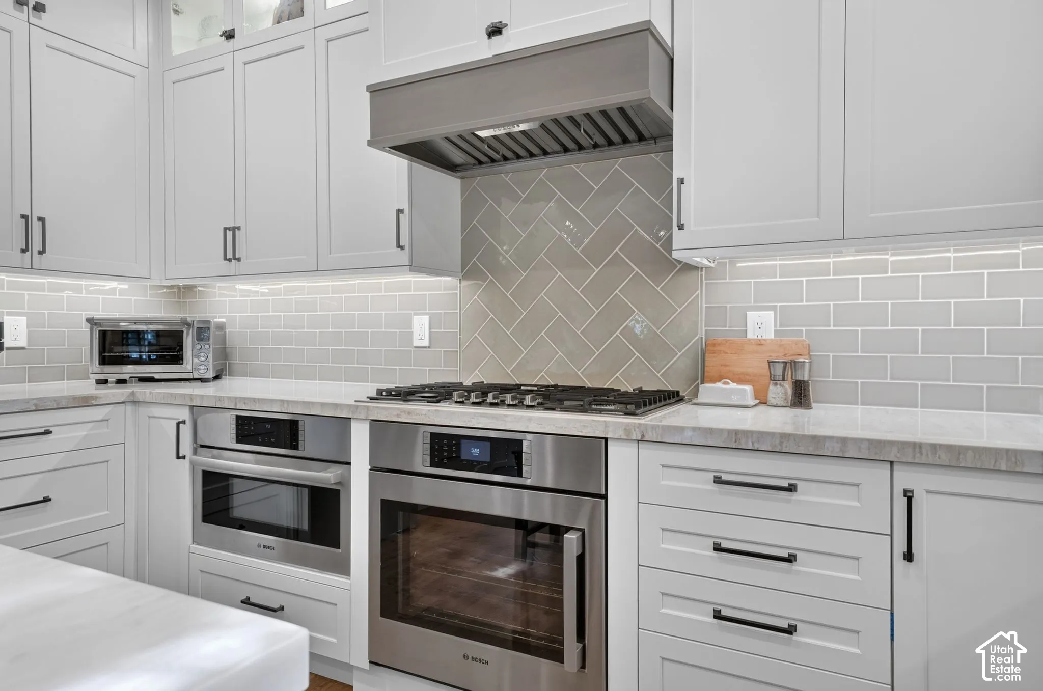 Kitchen featuring custom shaker-style cabinetry, herringbone-pattern tile backsplash, stone countertops, stainless steel built-in appliances, and matte black hardware