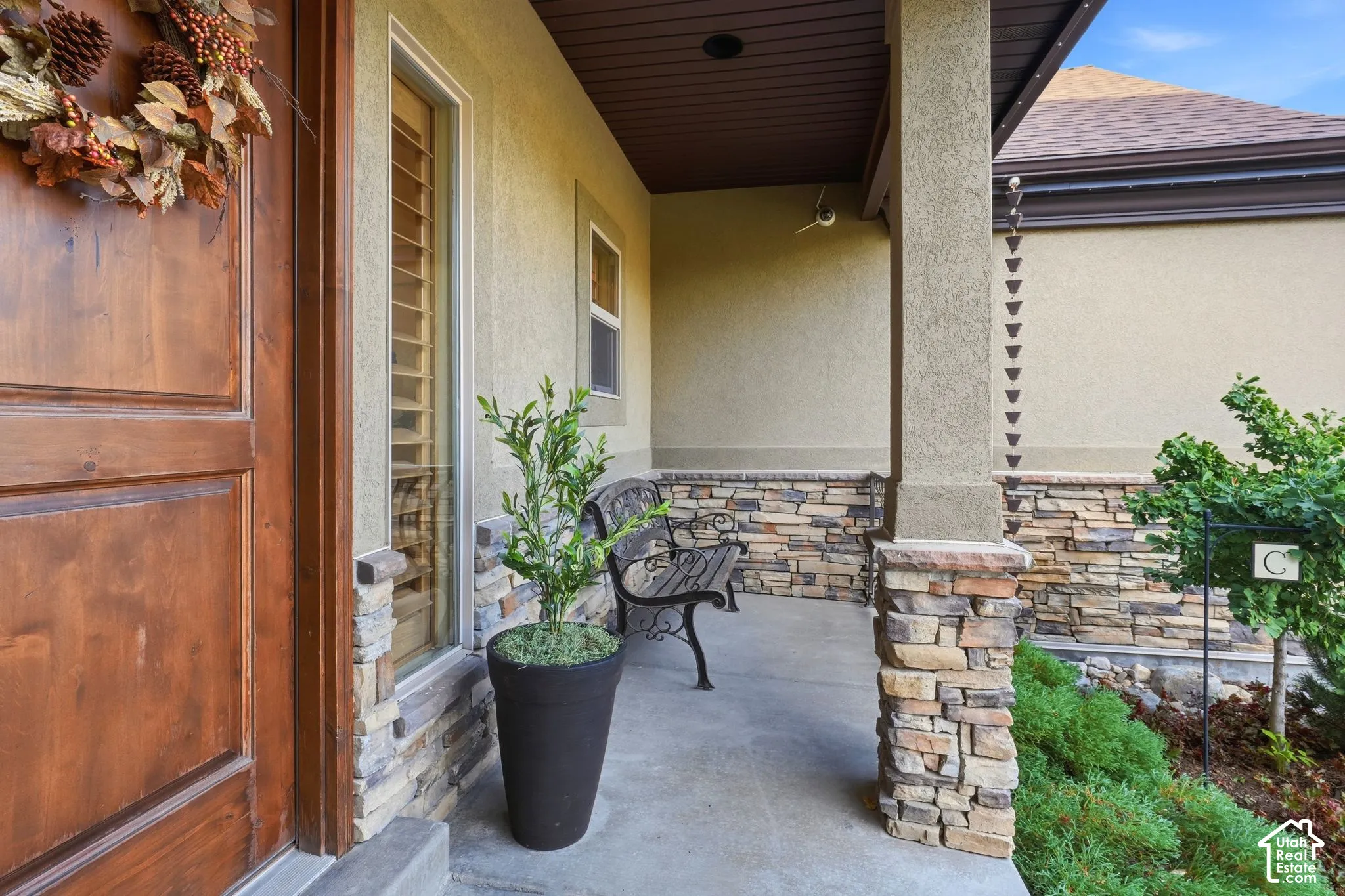 Covered entryway featuring a stained wood front door, natural stone veneer siding, concrete porch, and a metal rain chain