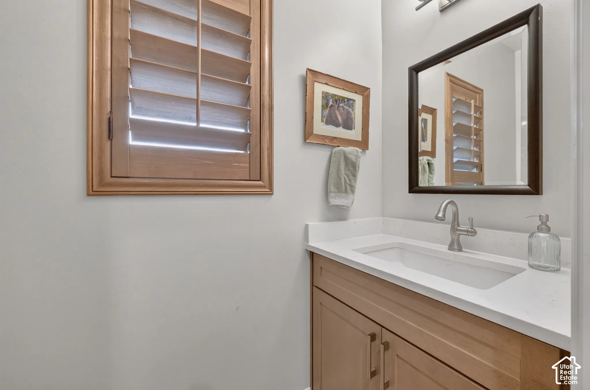 Bathroom featuring a light wood finish vanity, stone-composite countertop with an integrated rectangular sink, wall-mounted wood plantation shutters, and a framed mirror