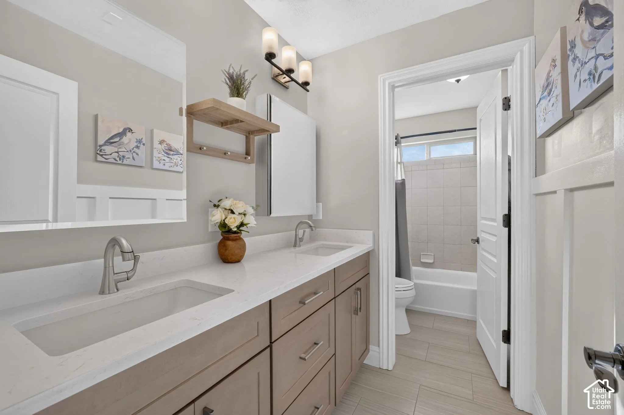 Dual-sink vanity featuring neutral-toned cabinetry, light-colored stone-finish countertops, three-light wall-mounted fixture, and tile flooring