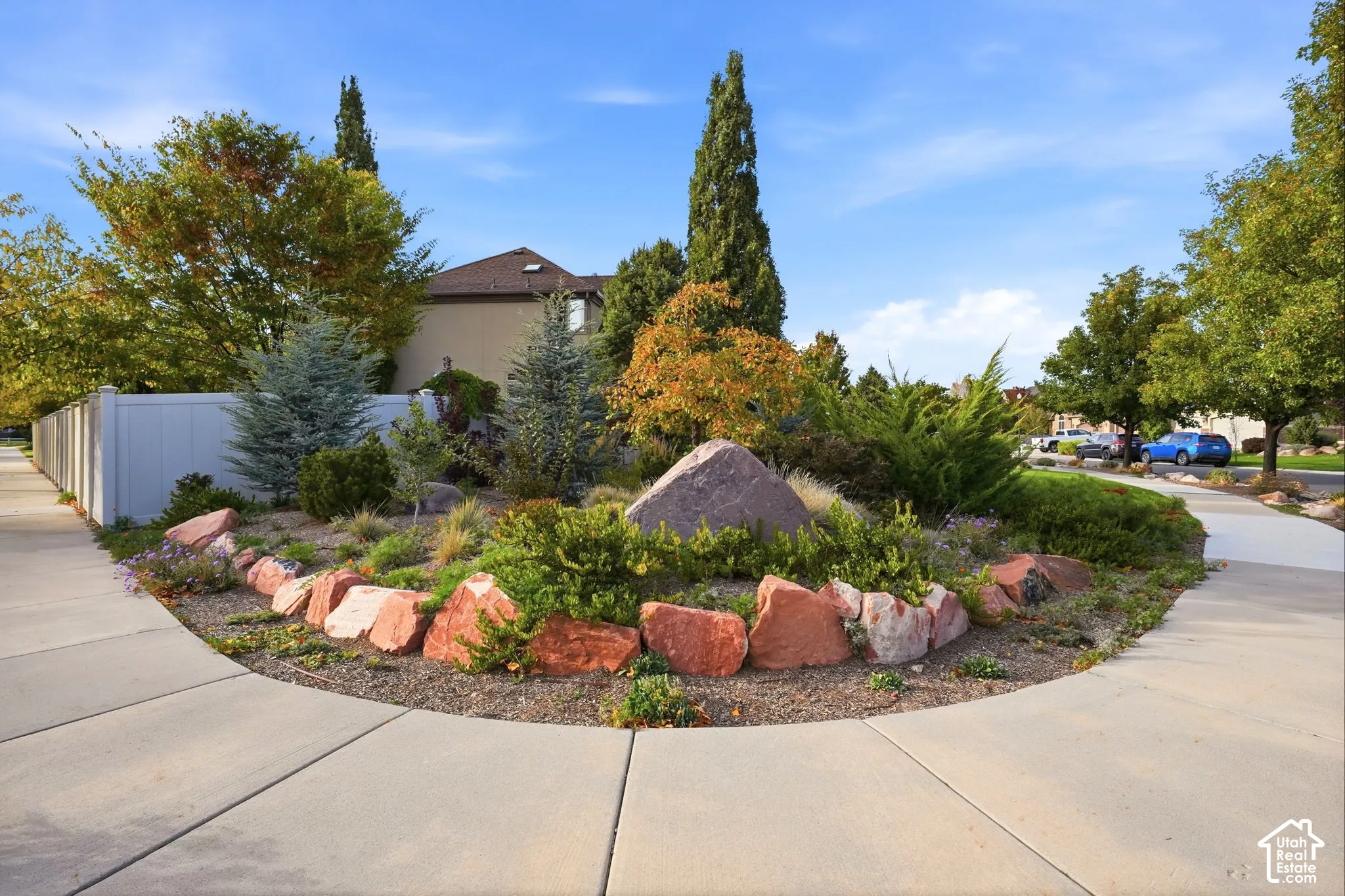 Landscaped corner lot featuring mature evergreen trees, ornamental shrubs, decorative rock mulch, and large natural stone border accents