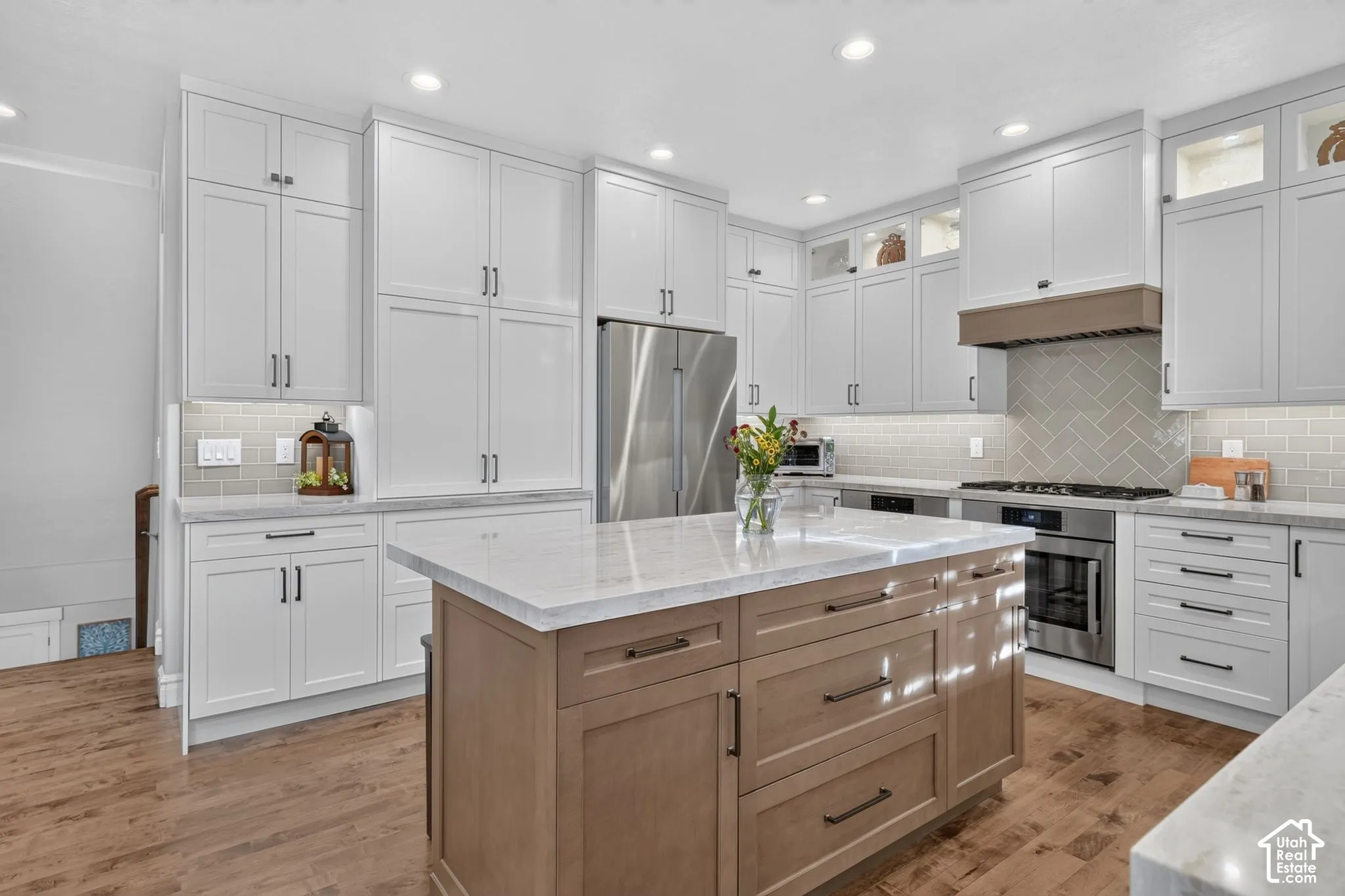 Kitchen featuring white shaker-style cabinetry, stone countertops, a wood-finish central island with drawers, herringbone-pattern tile backsplash, and wood-finish flooring