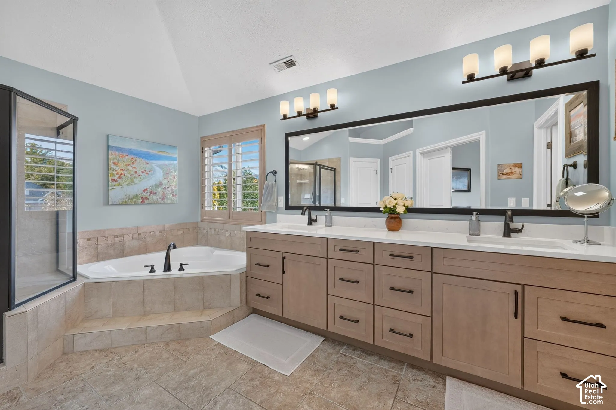 Dual-sink vanity with light-tone wood cabinetry, corner soaking tub with tile surround, walk-in glass-enclosed shower, expansive wall-mounted mirror, and plantation shutters