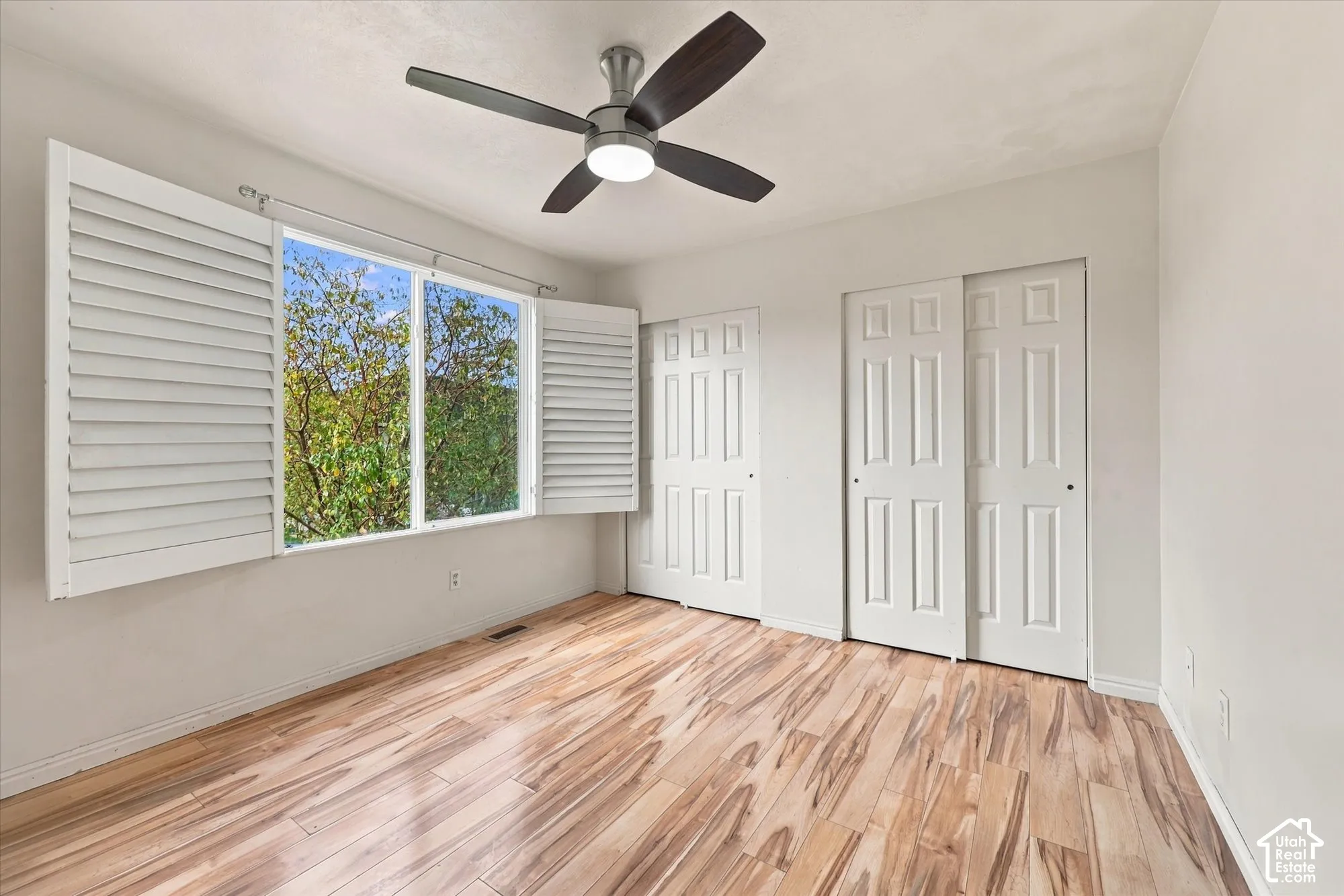 Room featuring wood-finish flooring, plantation shutters, dual-panel closet doors, and a ceiling-mounted light fixture with a fan