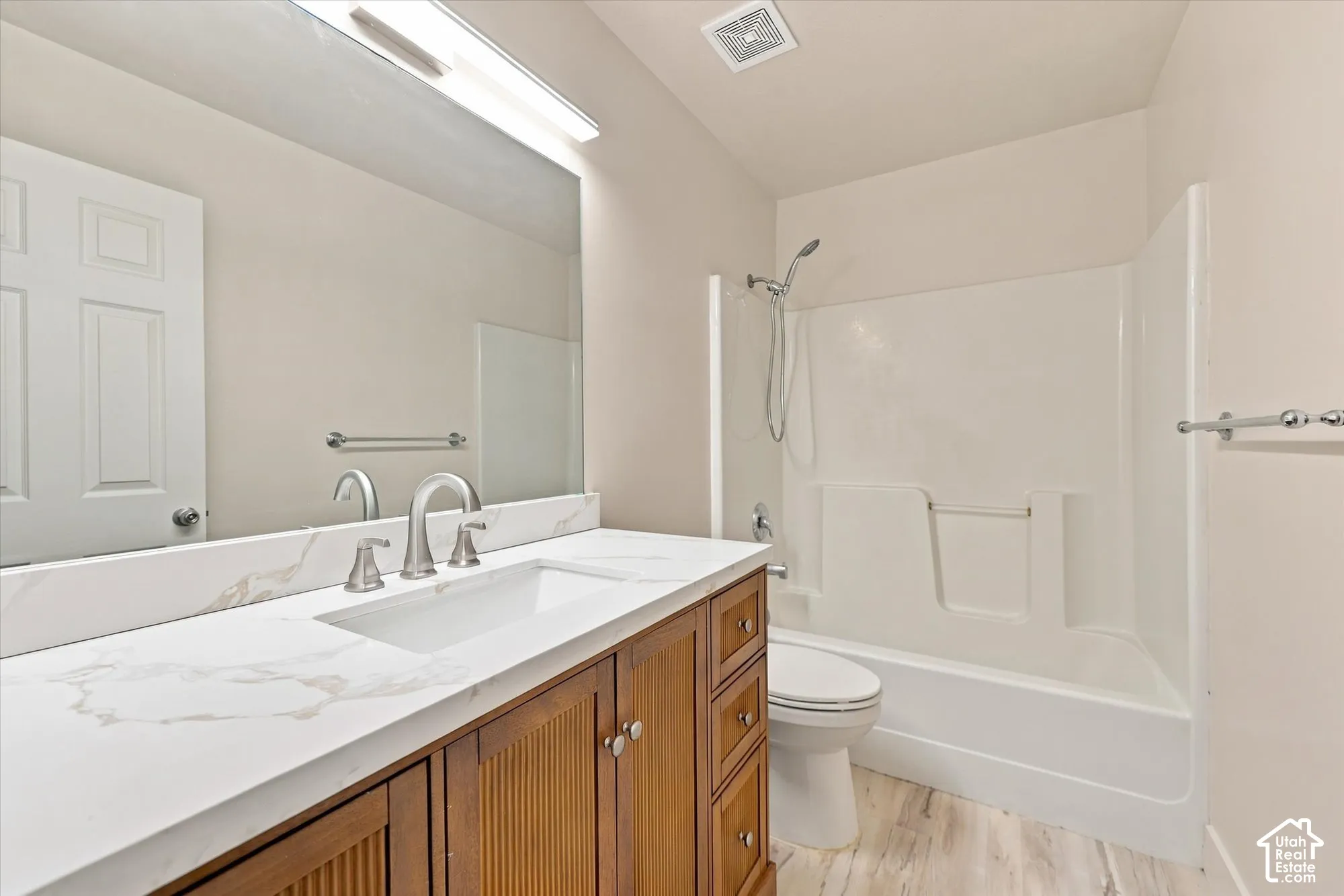 Bathroom featuring wood-finish vanity cabinetry, stone-look countertop with integrated rectangular sink, full-width mirror, brushed nickel faucet hardware, and a tub-shower combination with surround