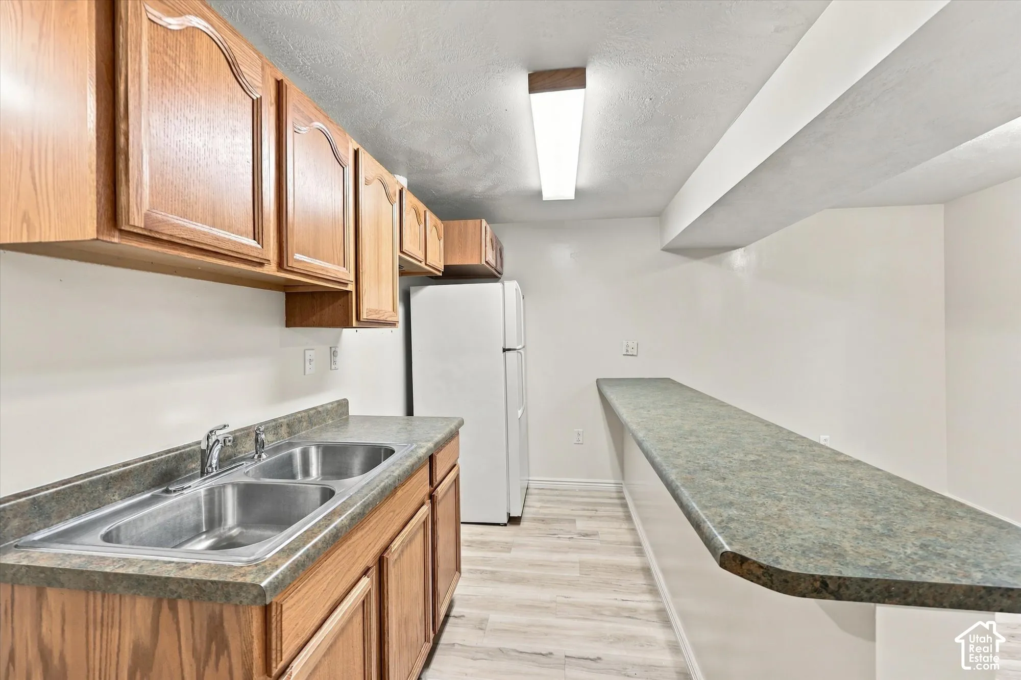 Kitchen area featuring light wood-finish cabinetry, dual-basin stainless steel sink, expansive solid-surface countertop, and wood-finish flooring