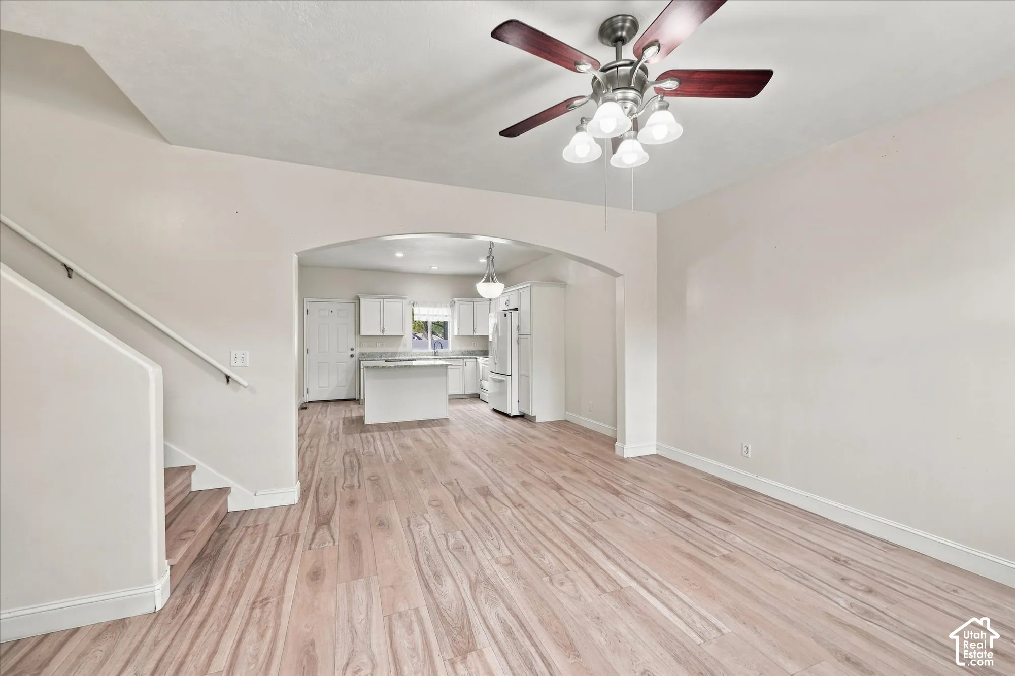 Open-concept interior featuring wood-finish flooring, a vaulted ceiling with a multi-light fan fixture, a prominent architectural archway, and a staircase with a white handrail