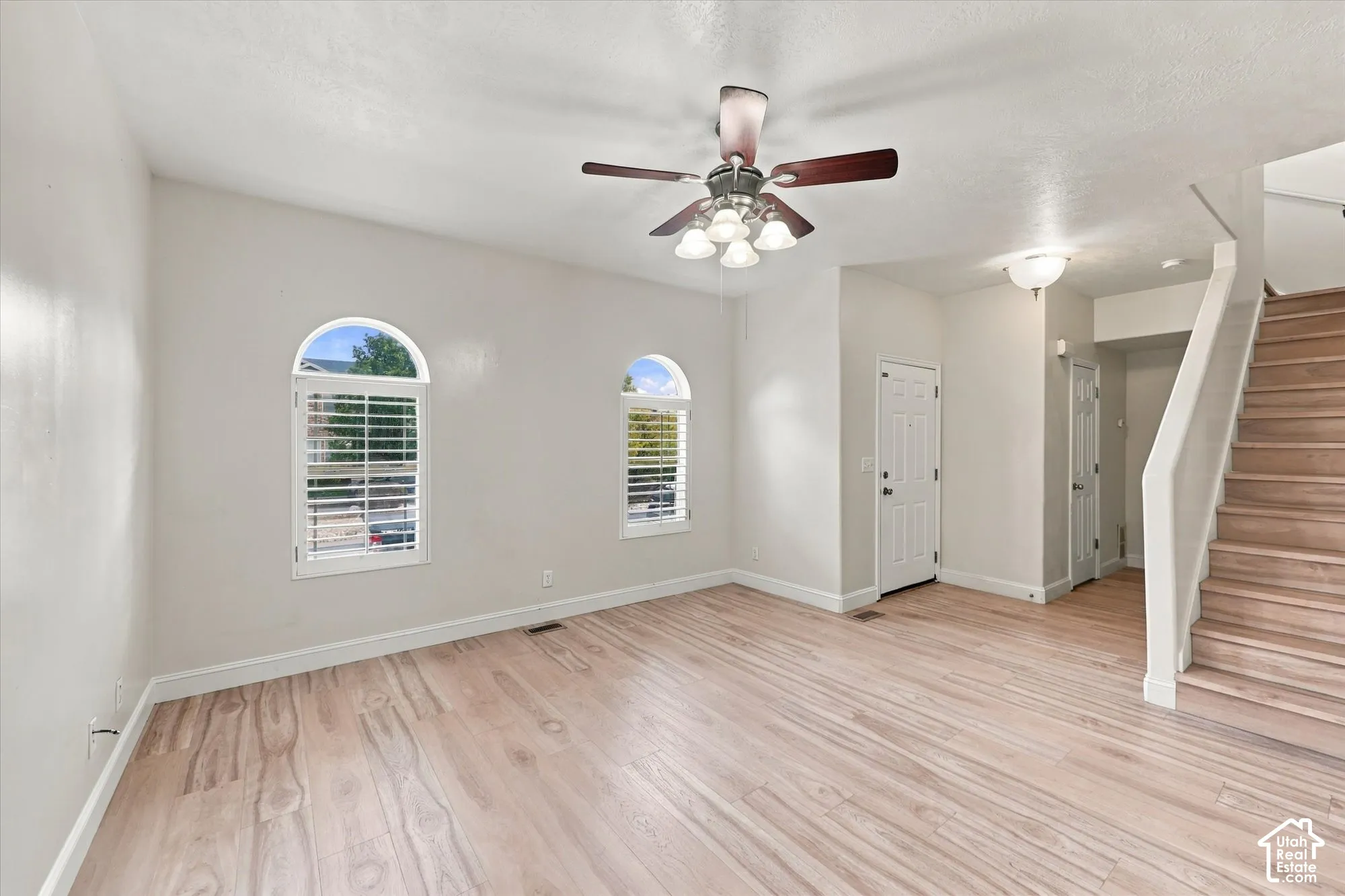 Interior space featuring wood-finish flooring, two arched windows with plantation shutters, a ceiling fan with multi-light fixture, and a staircase with wood-finish treads