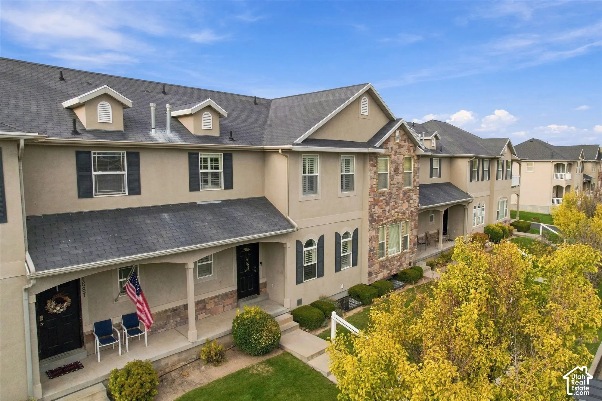 Multi-level exterior featuring tan stucco siding, stone veneer accents, dark-toned shutters, and a shingled roof with dormers