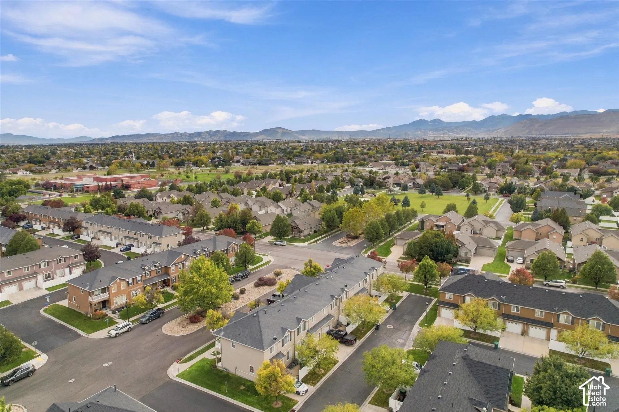 Aerial perspective featuring multi-level residential structures, asphalt roadways, mature deciduous trees, expansive manicured lawns, and a mountain range backdrop