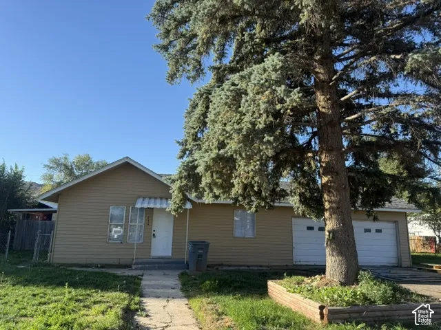 Single-story residence featuring horizontal siding, an attached two-car garage, concrete walkway, and mature evergreen tree within a raised landscape bed