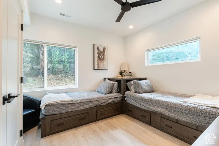 Built-in wood-finish bed frames with integrated drawer storage, recessed ceiling lighting, matte black ceiling fan, and large windows with natural forest exterior scenery