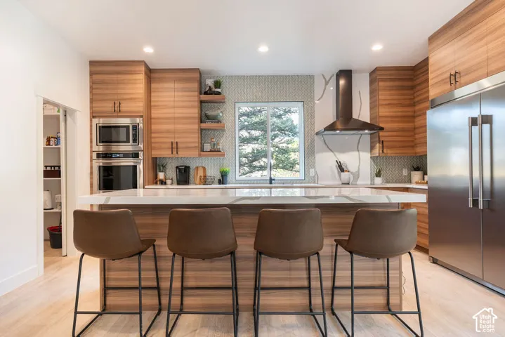 Kitchen featuring horizontal-grain wood cabinetry, a large waterfall-edge island with solid surface countertop, built-in stainless steel appliances, a geometric tile backsplash, and recessed lighting