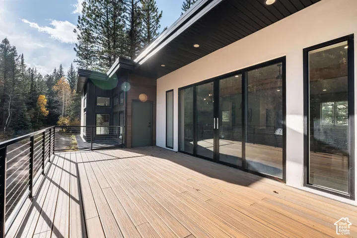 Expansive deck featuring wood-finish decking, black metal cable railing, oversized sliding glass doors, recessed lighting, and a modern smooth-stucco exterior facade