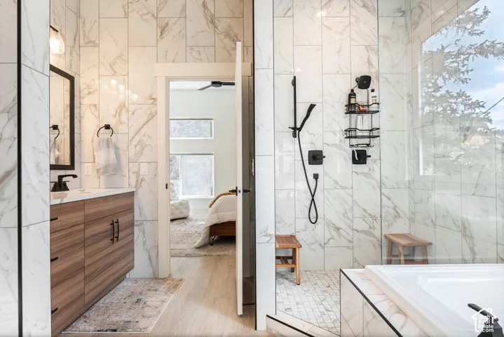 Bathroom featuring a wood-finish vanity with matte black hardware, floor-to-ceiling stone-patterned tile, glass-enclosed shower with a handheld fixture, and a built-in soaking tub