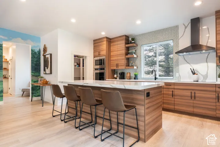 Kitchen featuring flat-panel wood-grain cabinetry, waterfall-edge quartz island, built-in stainless steel double oven, herringbone patterned tile backsplash, and matte black range hood