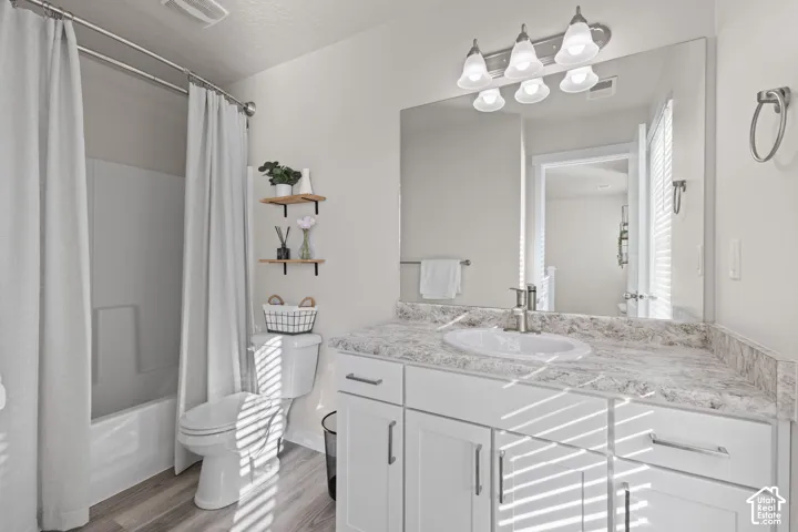 Bathroom featuring a white vanity cabinet with stone-look countertop, integrated sink, large wall-mounted mirror, four-light vanity fixture, and wood-finish flooring