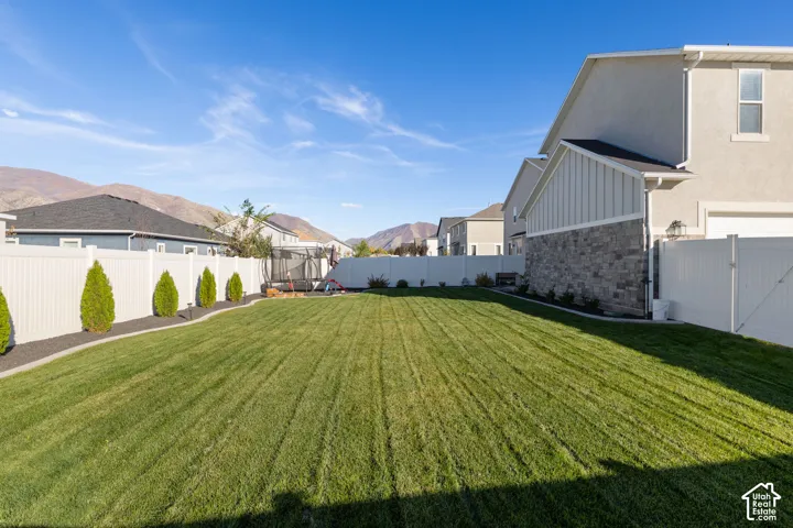 Fenced backyard featuring a manicured lawn, stone exterior accents, and mature ornamental shrubbery against a mountain backdrop