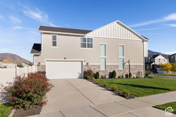 Two-story residence featuring light-toned stucco siding, stone veneer accents, vertical board-and-batten gable detailing, an attached garage, and a concrete driveway