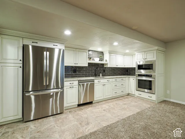 Kitchen downstairs featuring off-white cabinetry, stainless steel appliances, dark rectangular tile backsplash, and light-toned stone-patterned tile flooring