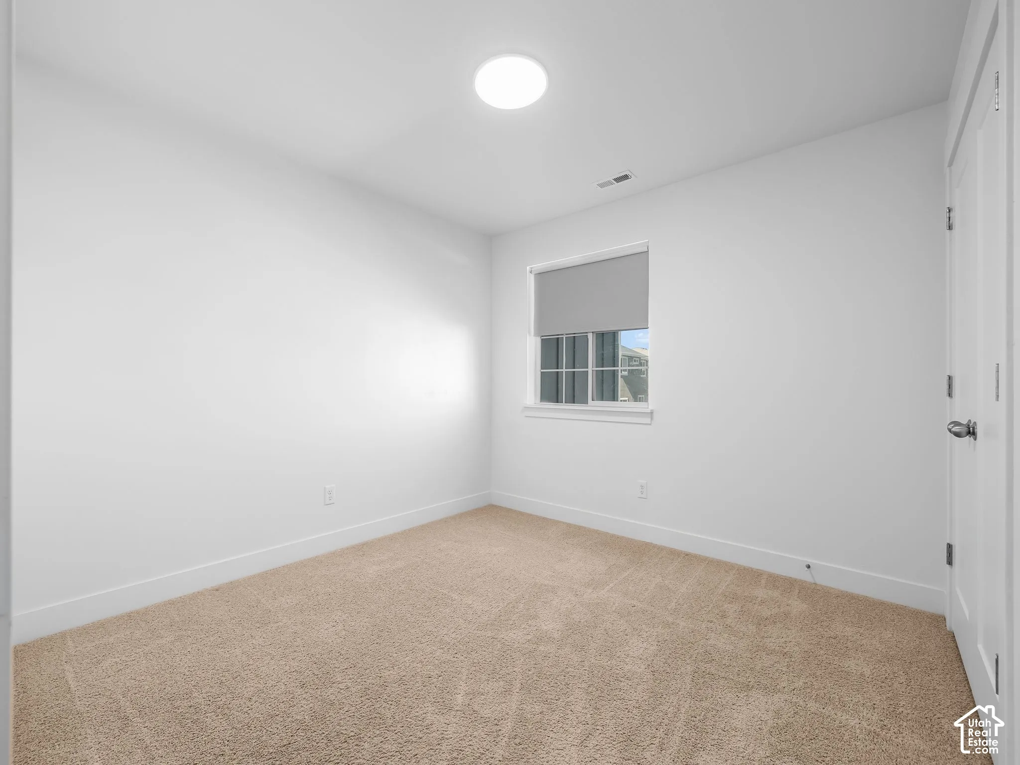 Neutral interior featuring neutral-toned carpeting, white walls, a flush-mount ceiling light fixture, and a window with a retractable shade