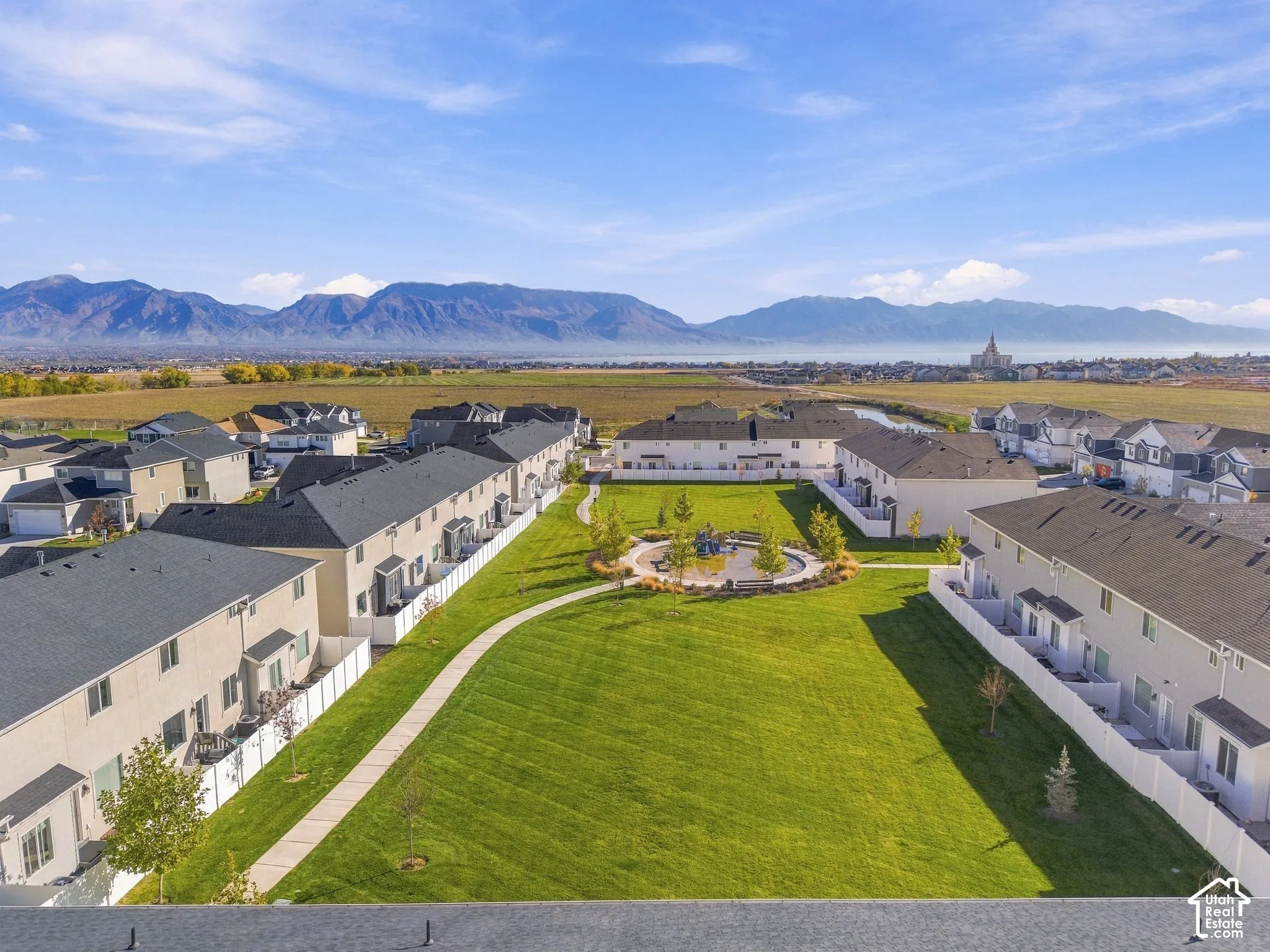 Aerial perspective of residential community featuring a central common green space, paved walkways, mountain range backdrop, and perimeter white vinyl fencing