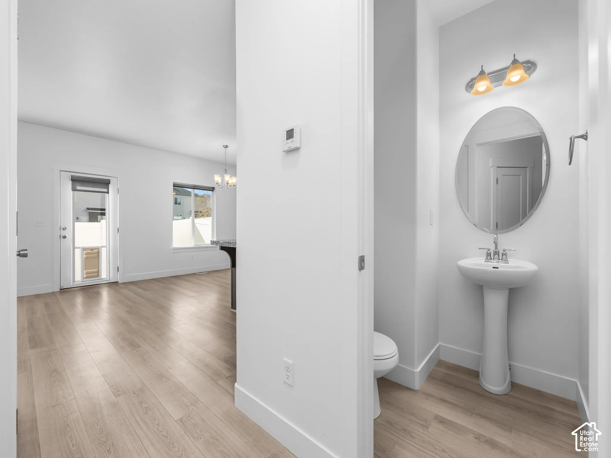 Powder room featuring a pedestal sink, oval wall-mounted mirror, and multi-bulb vanity light, complemented by wood-finish flooring extending into the main area