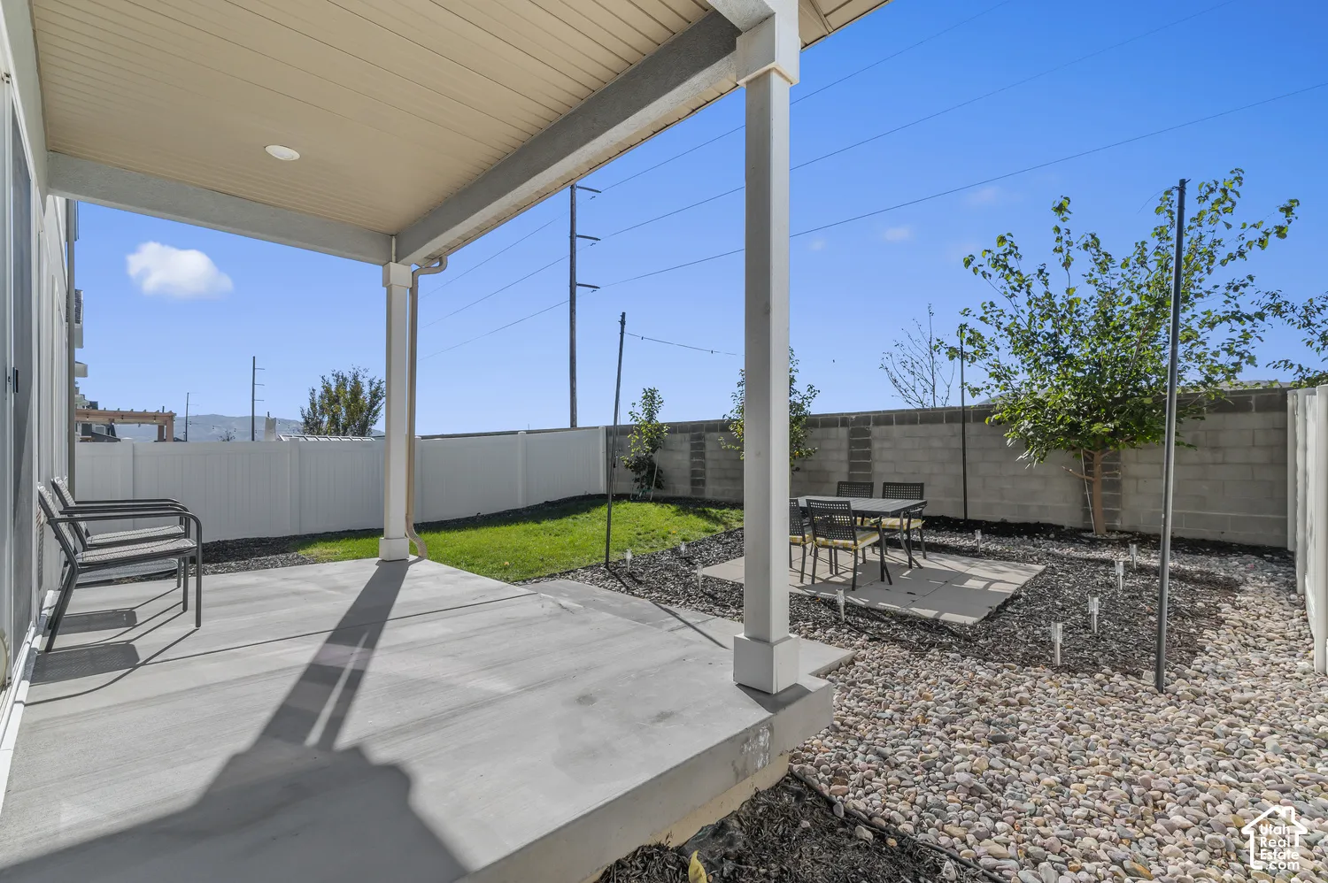 Covered concrete patio with overhead lighting, white vinyl perimeter fencing, stone-mulched yard, and manicured grass section