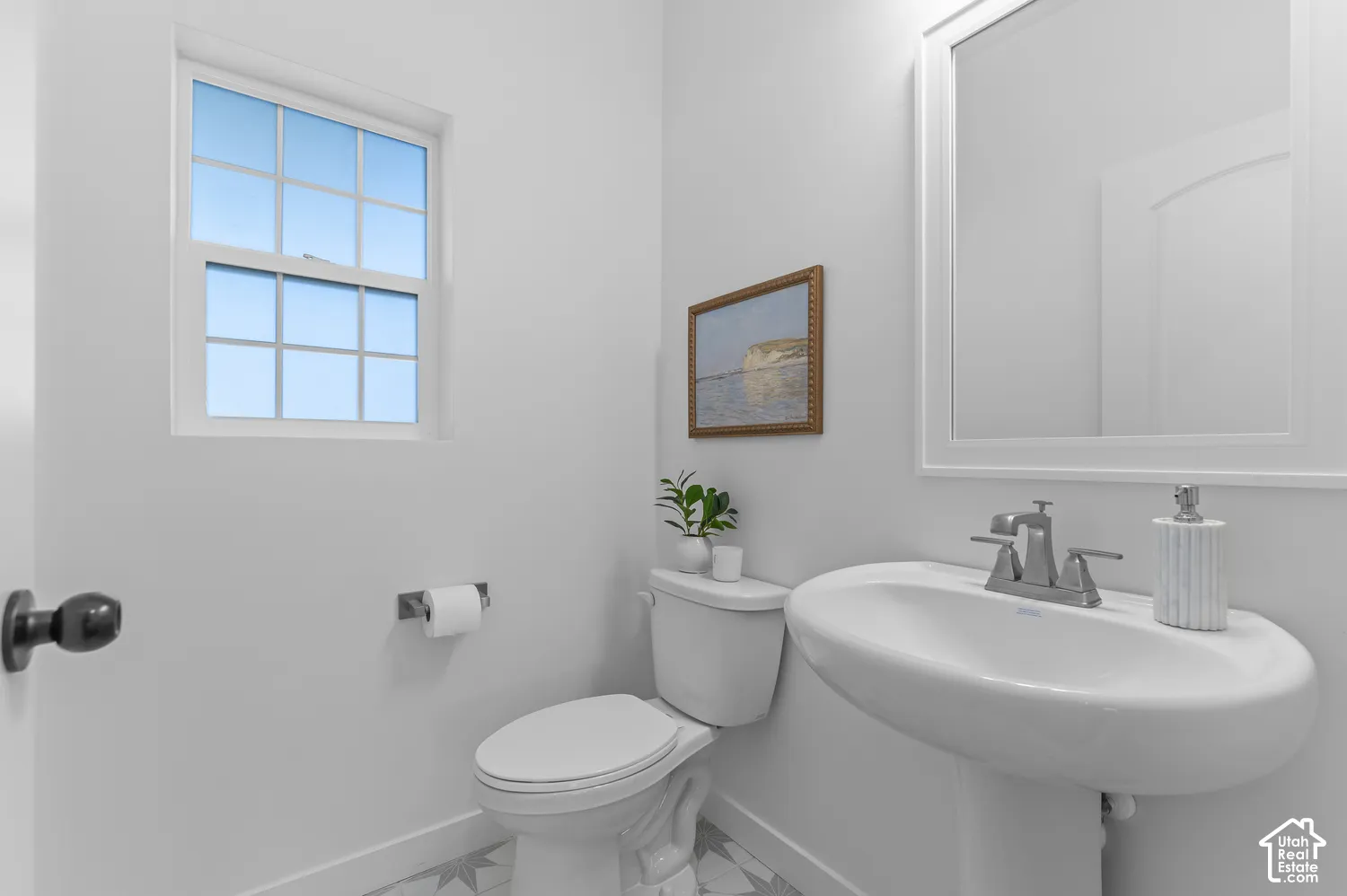 Bathroom featuring a white pedestal sink with a metallic faucet, a wall-mounted mirror with a white frame, a single-hung window with a grid pattern, and patterned tile flooring