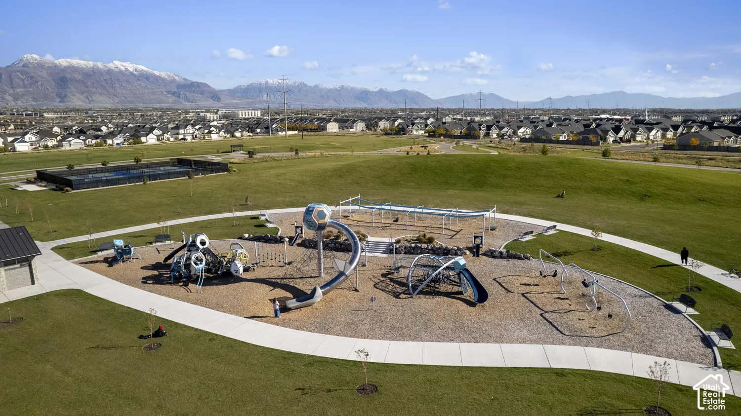 Expansive community park featuring playground equipment, a tennis court, manicured green space, concrete pathways, and a mountain backdrop