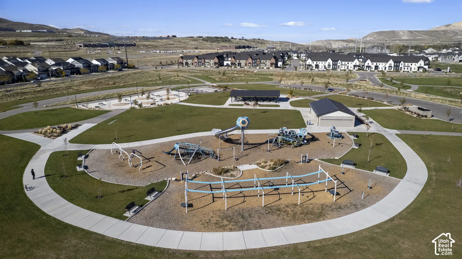Expansive community park featuring a playground, multi-purpose pavilion, concrete walking paths, manicured green space, and mountain backdrop