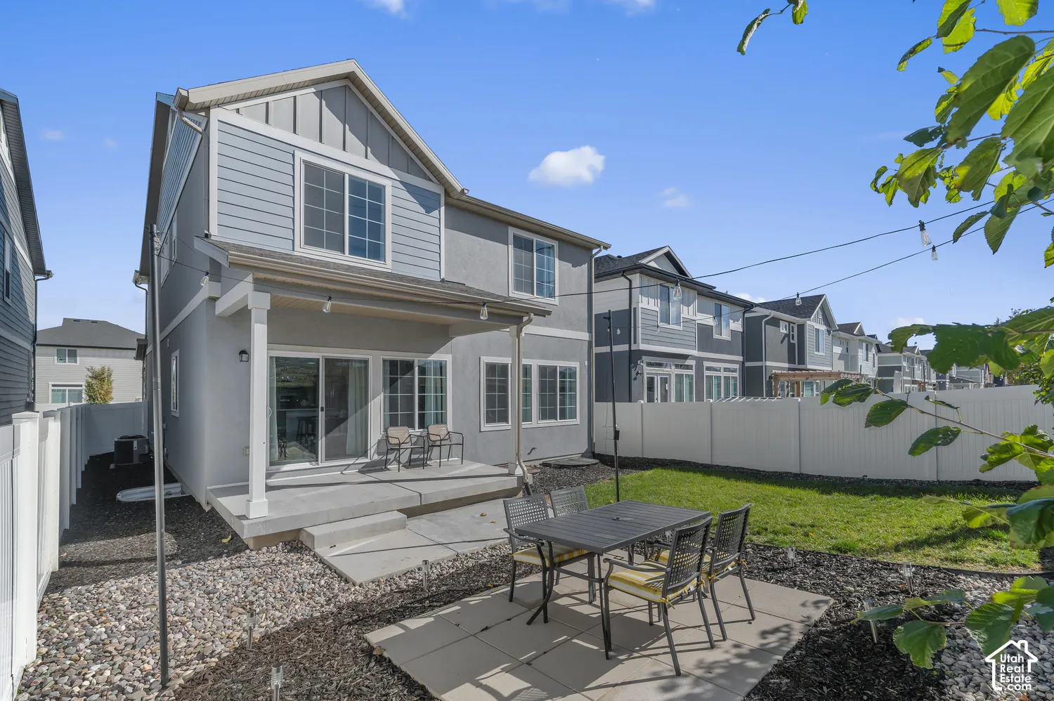 Two-story exterior featuring fiber cement siding, a covered concrete patio, a secondary stone paver patio, and white vinyl perimeter fencing