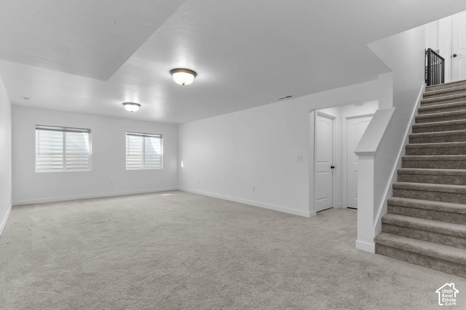 Expansive interior area featuring wall-to-wall carpeting, dual flush-mount ceiling lights, two windows with horizontal blinds, and carpeted stairs with a metal balustrade