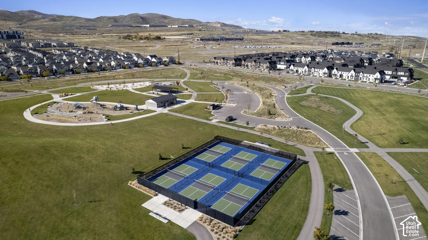 Aerial perspective of a multi-court pickleball facility with perimeter fencing, paved pedestrian pathways, manicured green space, and mountain backdrop