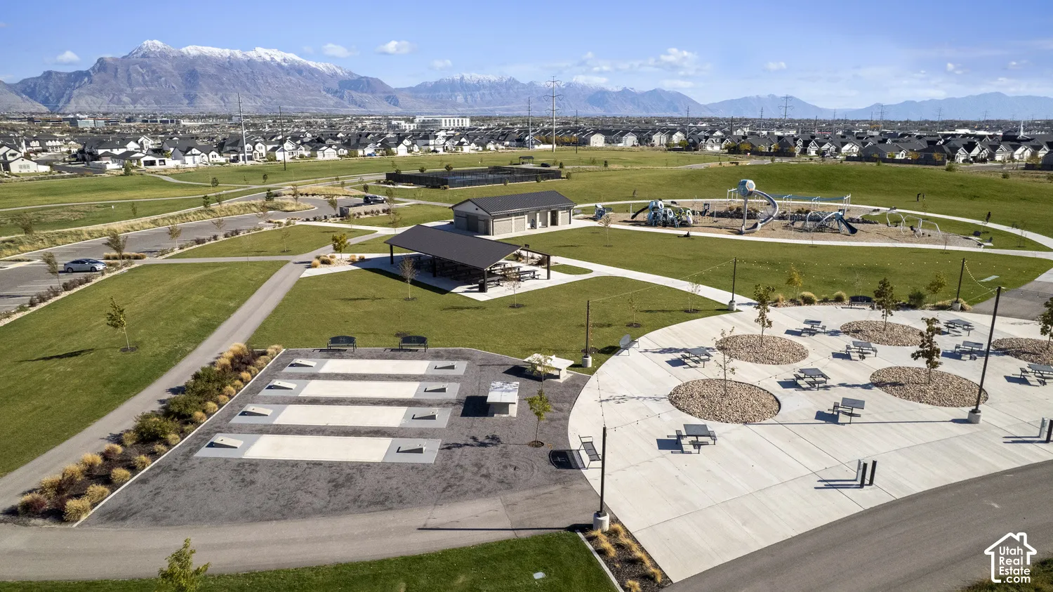 Community park area featuring a covered pavilion, playground equipment, concrete pathways, and recreational courts set against a mountain backdrop