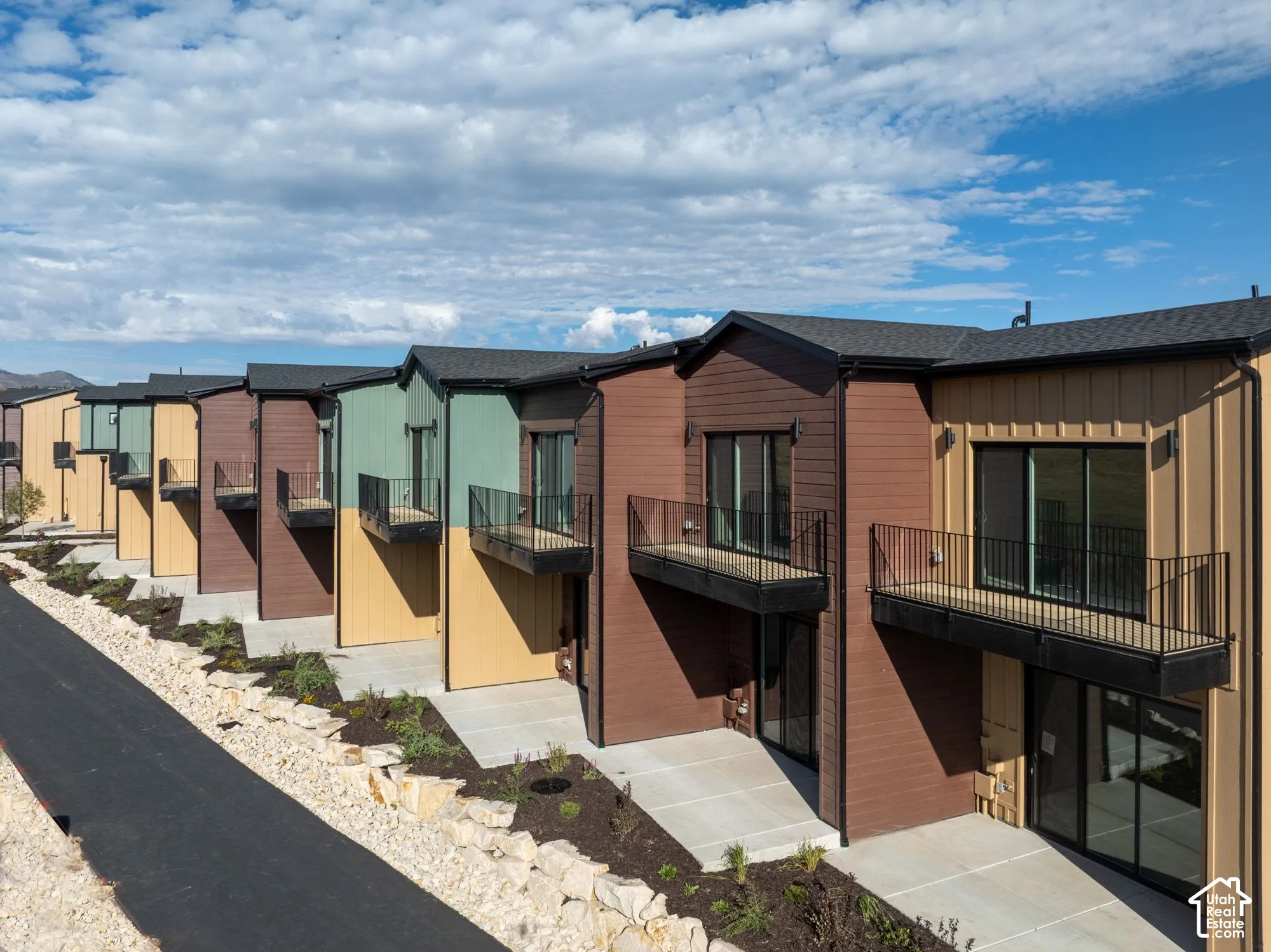 Multi-unit exterior featuring mixed-material siding in earth tones, private elevated balconies with metal railings, sliding glass doors, and concrete patios