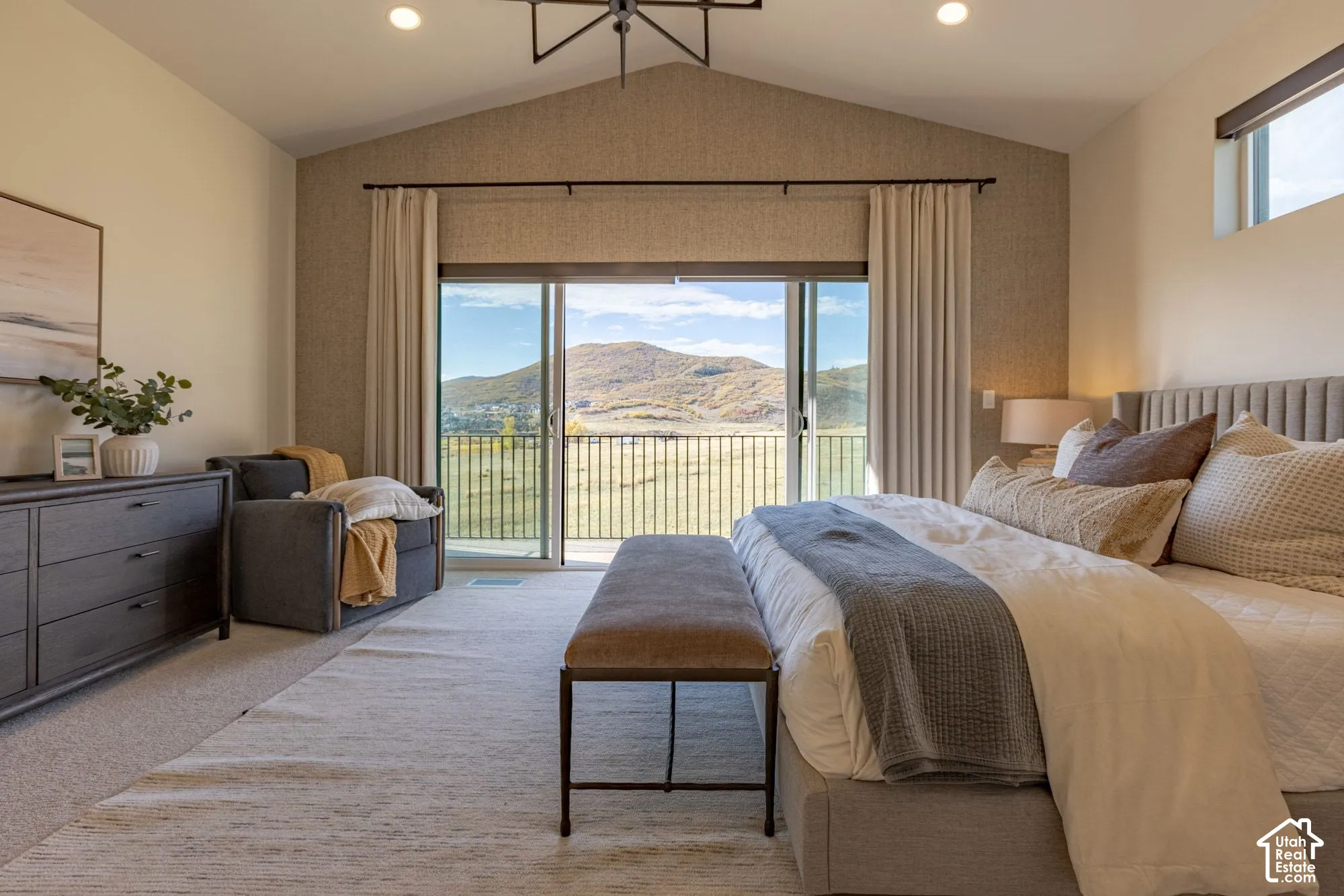 Vaulted ceiling with a geometric chandelier, textured accent wall, sliding glass doors opening to a balcony with metal railing, and expansive mountain scenery