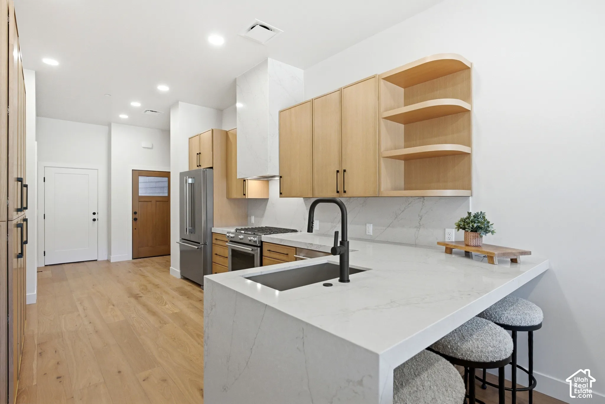 Kitchen featuring light wood-finish cabinetry, waterfall-edge stone countertop with a matte black faucet, integrated stone backsplash, recessed lighting, and wood-finish flooring