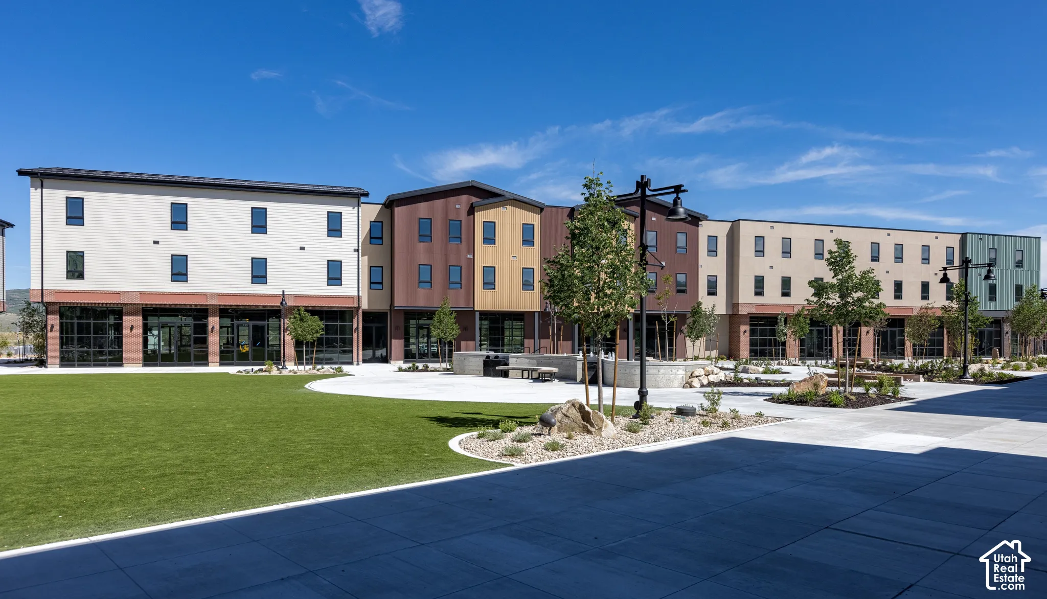 Multi-story architectural exterior featuring metal and paneled siding, expansive ground-level glass storefronts, manicured lawn, stone hardscaping, and decorative street lamps