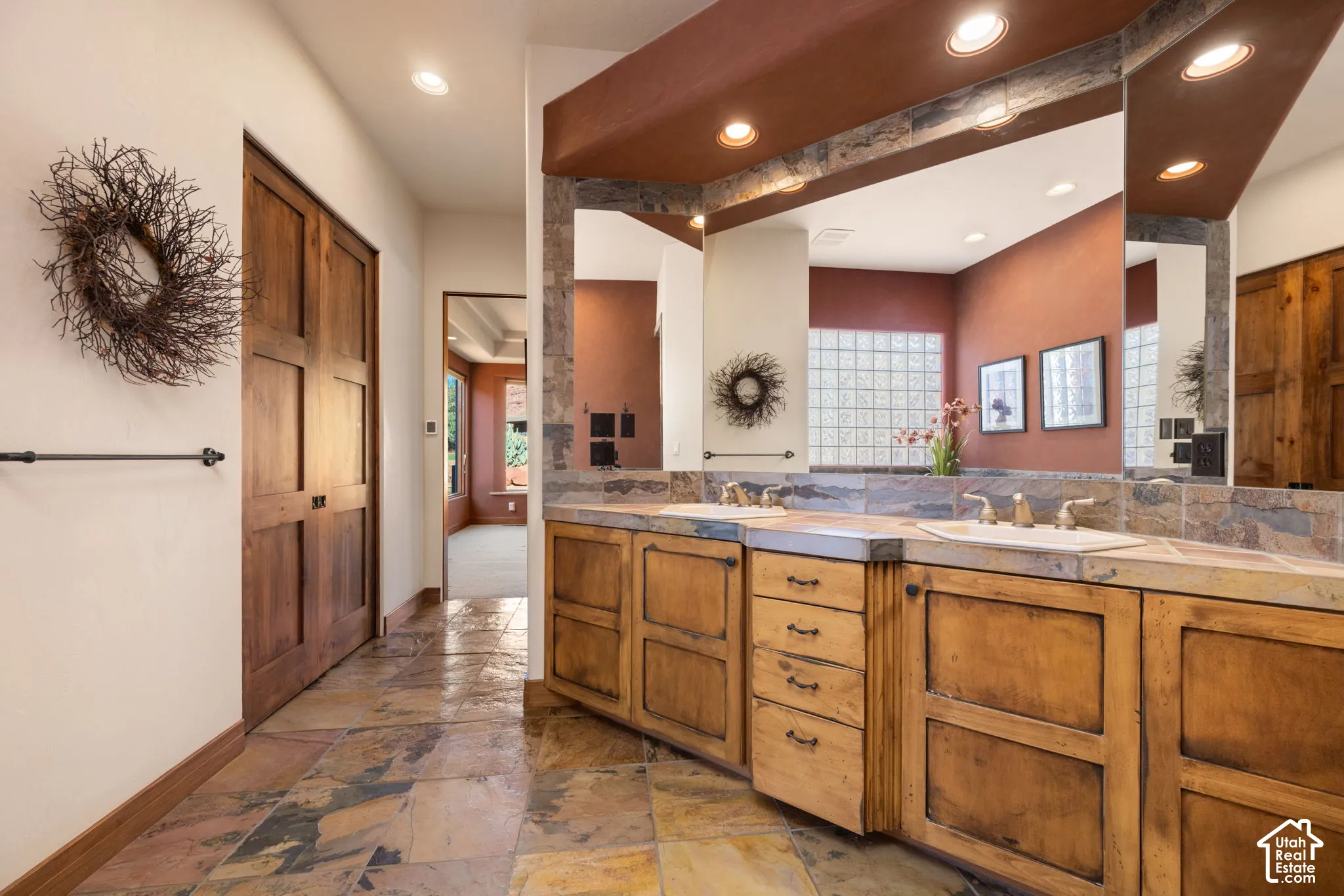 Dual-vanity cabinetry with natural wood finish, multi-tonal stone tile flooring, stone countertop with integrated sinks, recessed ceiling lighting, and large-format wall mirrors with stone tile backsplash